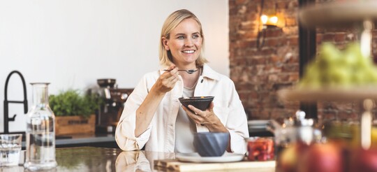 Frau sitzt lächelnd am Tisch und isst aus einer Schüssel mit Löffel.