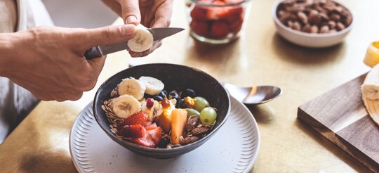 Frau schneidet Banane über einer Schüssel mit frischem Obst und Nüssen.