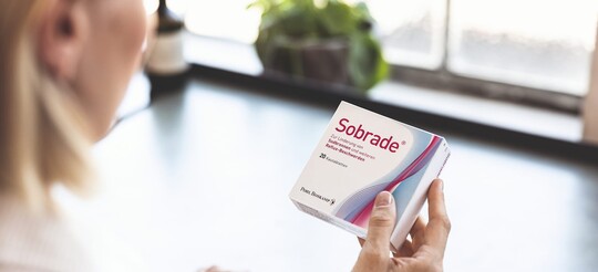 Blonde Frau ist von hinten zu sehen, wie sie eine Sobrade Verpackung mit 20 Kautabletten in der Hand hält und sich die Beschriftung durch liest
