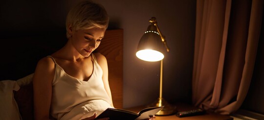 Eine Frau sitzt bei gedimmtem Licht lesend in ihrem Bett.