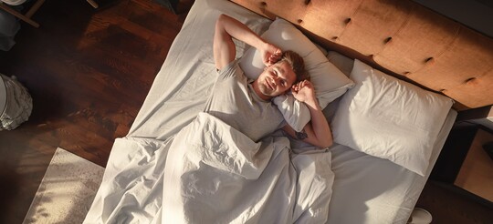 Ein Mann liegt am Morgen entspannt in seinem Bett und reckt die Arme an den Kopf.