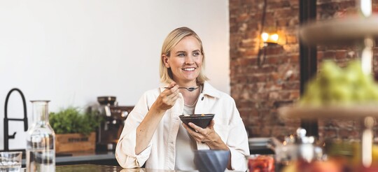 Frau sitzt lächelnd am Tisch und isst aus einer Schüssel mit Löffel.