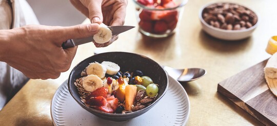 Frau schneidet Banane über einer Schüssel mit frischem Obst und Nüssen.
