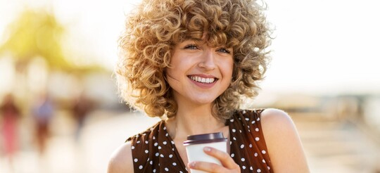 Eine Frau mit lockigen Haaren lächelt im Sonnenlicht in die Kamera. In der Hand trägt sie einen Kaffeebecher.