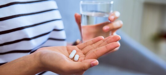 Eine Person hält zwei Tabletten und ein Glas Wasser zum Schlucken der Tabletten in den Händen.