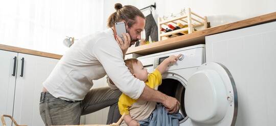 Ein telefonierender Vater kniet in der Küche vor einer Waschmaschine, während sein Baby an den Reglern spielt.