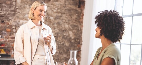 Zwei lächelnde Frauen unterhalten sich in gemütlicher Atmosphäre, eine hält ein Glas Wasser in der Hand.