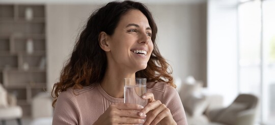 Lächelnde Frau mit welligem braunen Haar hält ein Glas Wasser in der Hand und schaut zur Seite.