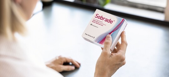 Blonde Frau ist von hinten zu sehen, wie sie eine Sobrade Verpackung mit 20 Kautabletten in der Hand hält und sich die Beschriftung durch liest
