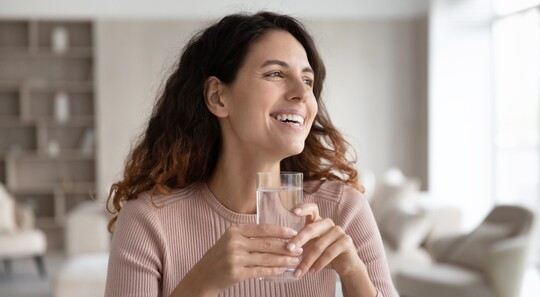 Lächelnde Frau mit welligem braunen Haar hält ein Glas Wasser in der Hand und schaut zur Seite.