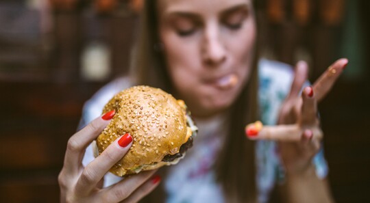 Nahaufnahme einer Frau mit Burger in der Hand, während sie sich die Finger ableckt.