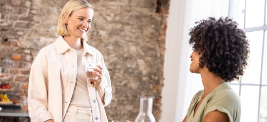 Zwei lächelnde Frauen unterhalten sich in gemütlicher Atmosphäre, eine hält ein Glas Wasser in der Hand.