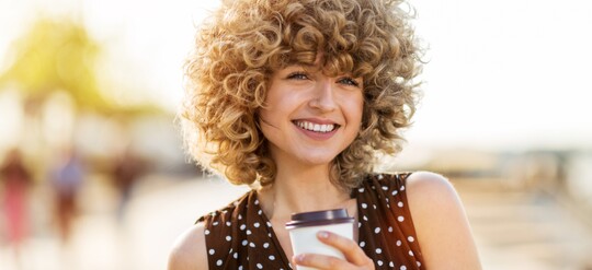 Eine Frau mit lockigen Haaren lächelt im Sonnenlicht in die Kamera. In der Hand trägt sie einen Kaffeebecher.
