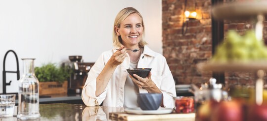 Frau sitzt lächelnd am Tisch und isst aus einer Schüssel mit Löffel.