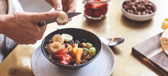 Frau schneidet Banane über einer Schüssel mit frischem Obst und Nüssen.