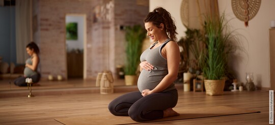 Eine schwangere Frau kniet auf einer Yogamatte und hält lächelnd ihren Bauch.