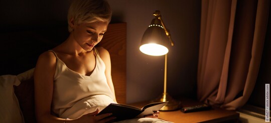 Eine Frau sitzt bei gedimmtem Licht lesend in ihrem Bett.