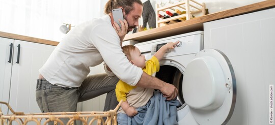 Ein telefonierender Vater kniet in der Küche vor einer Waschmaschine, während sein Baby an den Reglern spielt.