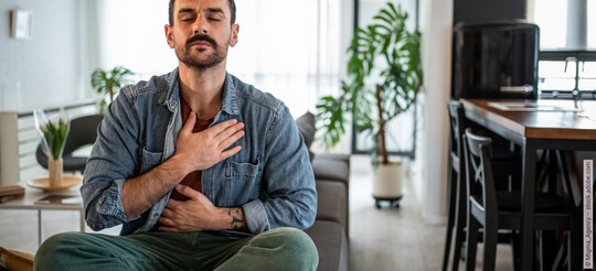 Ein Mann sitzt geschlossenen Augen auf einem Sofa und hält sich eine Hand auf die Brust und die andere auf den Magen.