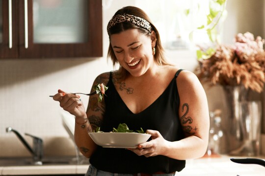 Eine lachende Frau mit Tattoos und einem Leopard-Haarband isst lächelnd einen frischen Salat.