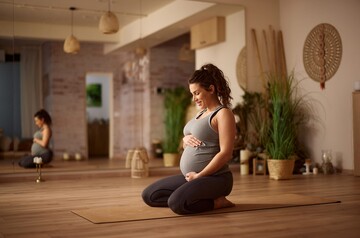 Eine schwangere Frau kniet auf einer Yogamatte und hält lächelnd ihren Bauch.