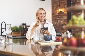 Frau sitzt lächelnd am Tisch und isst aus einer Schüssel mit Löffel.
